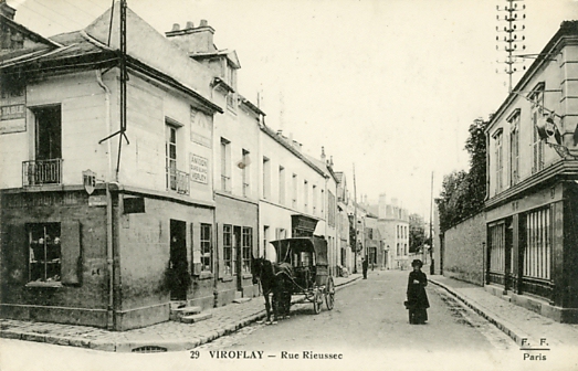 Belle perspective sur la rue rieussec  partir de la place de l’glise. Une voiture  cheval stationne devant le dbit de vins - picerie.